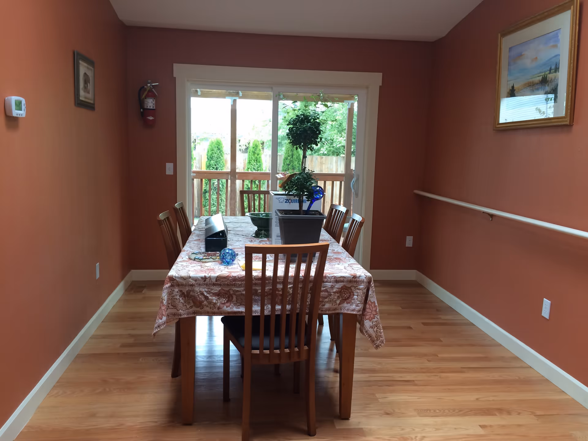A dining room with a wooden table covered by a floral tablecloth and surrounded by six wooden chairs. On the table, there is a potted plant, a blue glass decorative item, and some other small objects. The room has orange walls, a wooden floor, a handrail on the right wall, and a large sliding glass door at the back leading to an outdoor area with greenery.