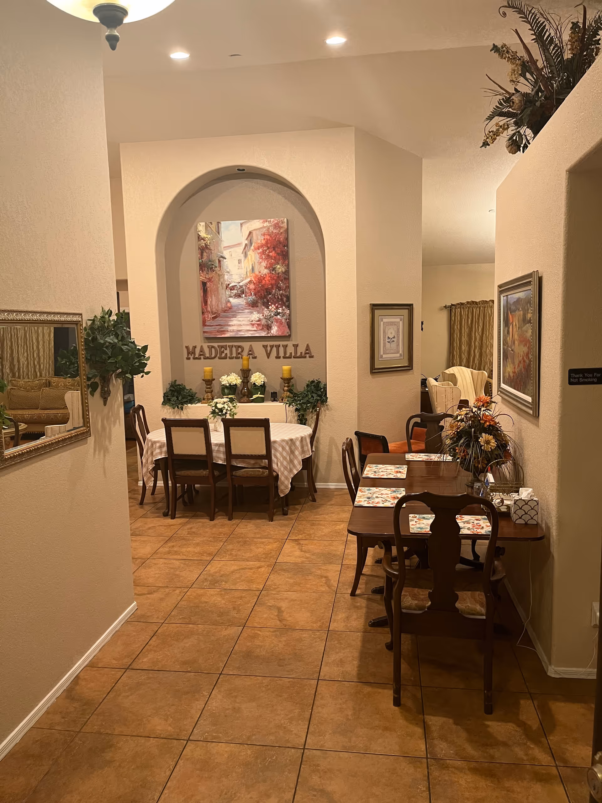 Interior view of a dining area at Madeira Villa Assisted Living featuring two wooden dining tables with chairs. One table is covered with a checkered tablecloth and decorated with candles and flowers, positioned under an archway with a painting and the words 'MADEIRA VILLA' on the wall. The other table has placemats and a floral centerpiece. The room has tiled floors, beige walls, framed artwork, and decorative plants.