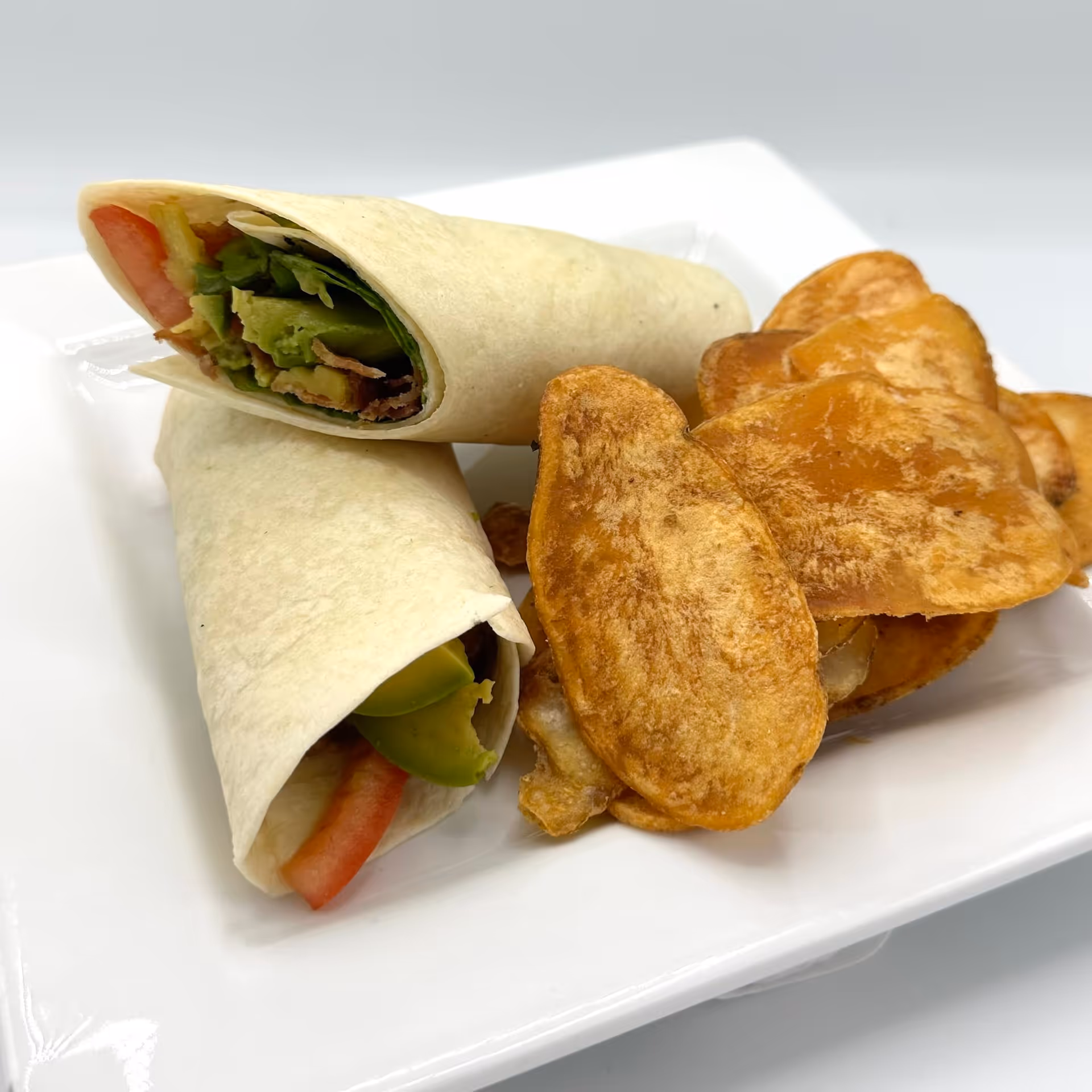 A white square plate with two tortilla wraps filled with lettuce, tomato, and avocado, served alongside a pile of thick-cut potato chips.