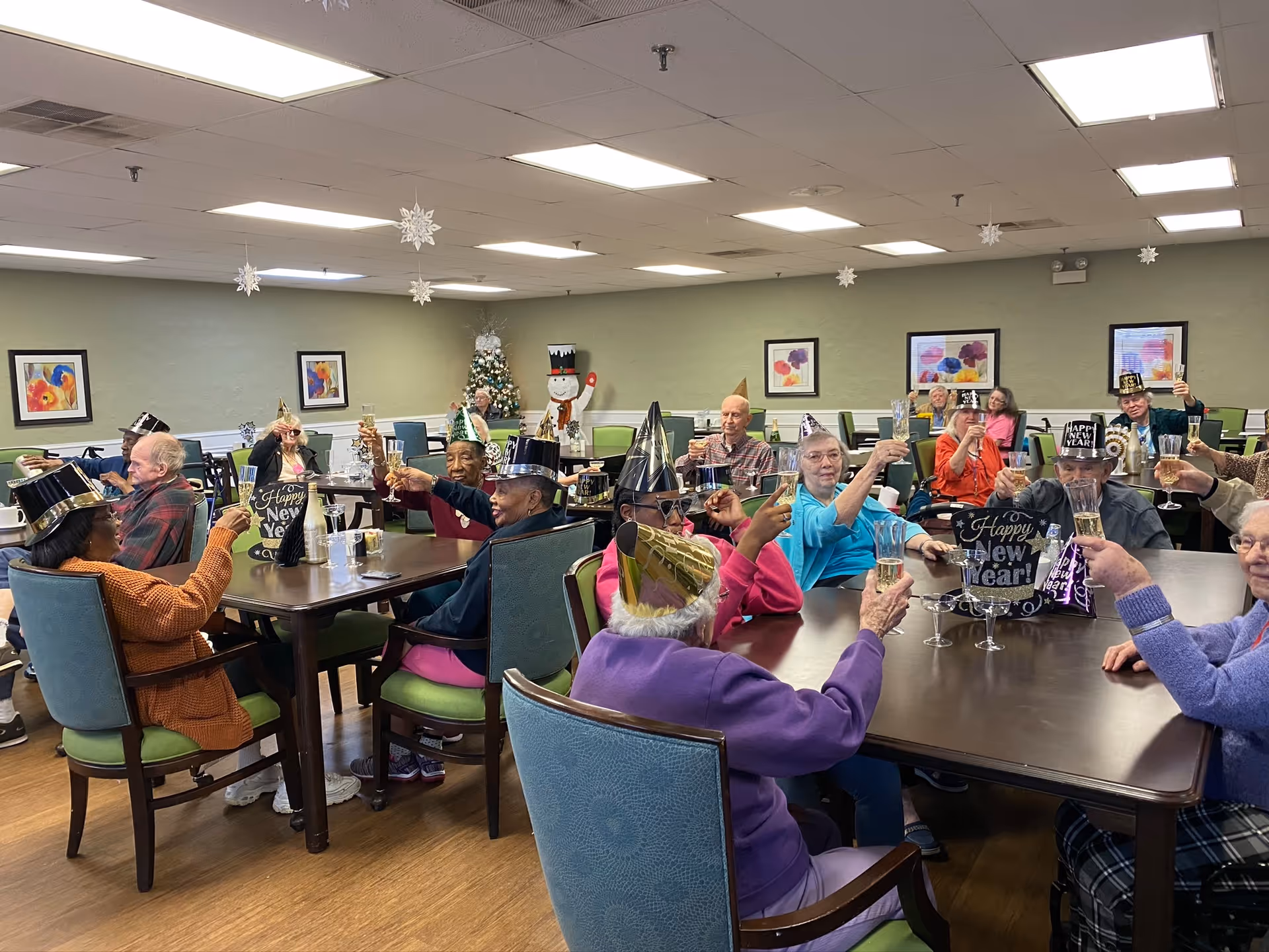 Residents in a senior living dining room raise glasses and wear party hats celebrating New Year.