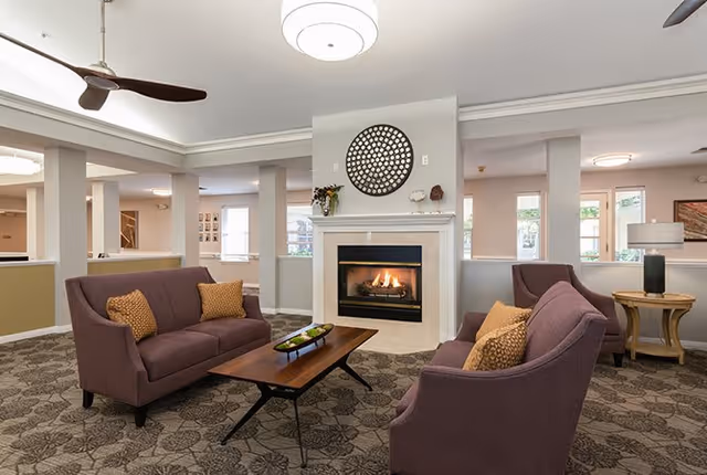 A cozy senior living facility common area with two purple sofas and a matching armchair arranged around a wooden coffee table. A fireplace with a decorative circular wall art above it is centered in the room. The space has patterned carpet flooring, neutral-colored walls, and large windows allowing natural light to enter. A ceiling fan and a modern ceiling light fixture are visible.