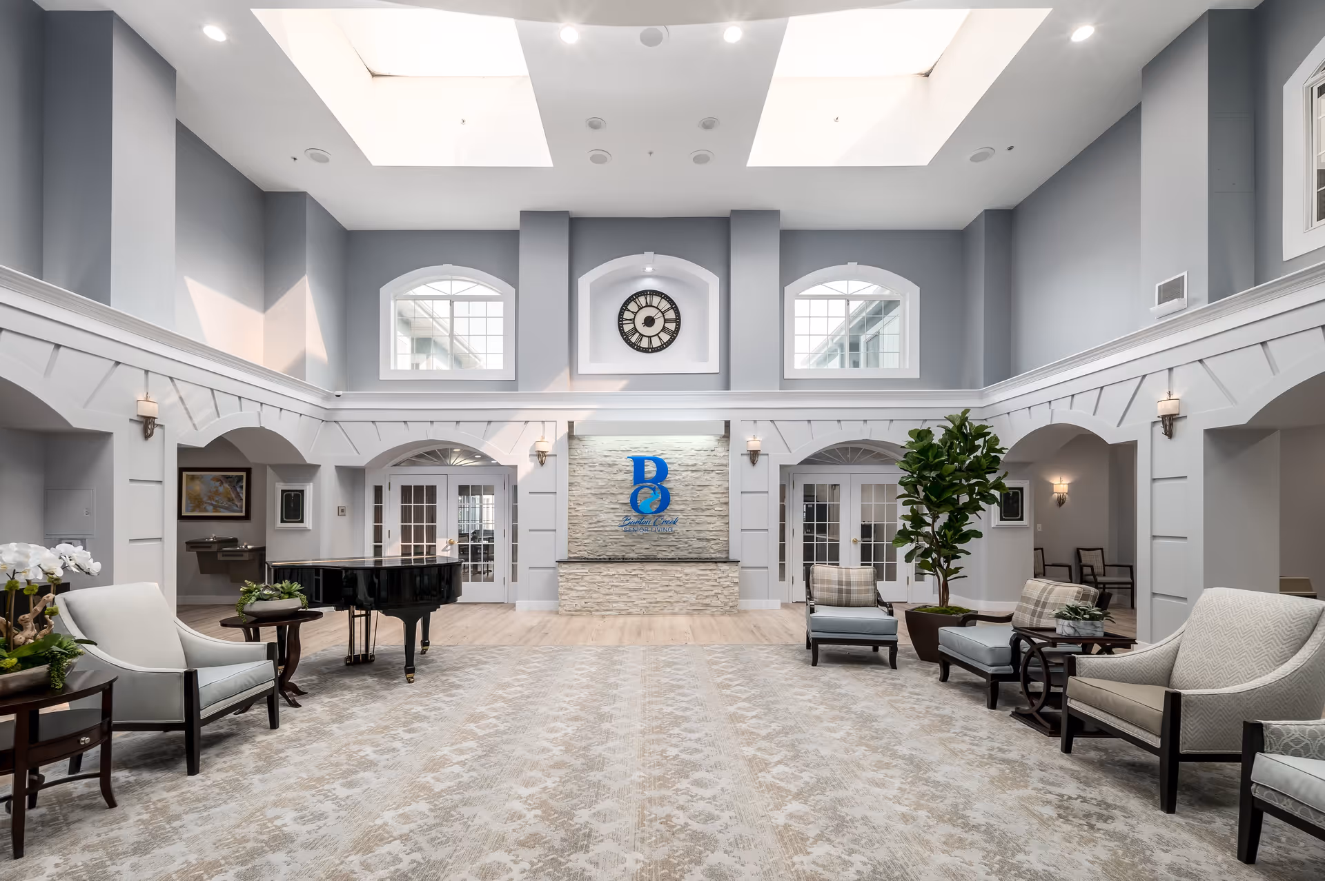 Spacious and elegant senior living facility common area with high ceilings and skylights. The room features comfortable seating with armchairs and small tables, a black grand piano, a large clock mounted above a stone accent wall with the blue Barton Creek Senior Living logo, and large windows letting in natural light. The walls are painted light gray with white trim, and there are decorative wall sconces and potted plants adding to the welcoming atmosphere.