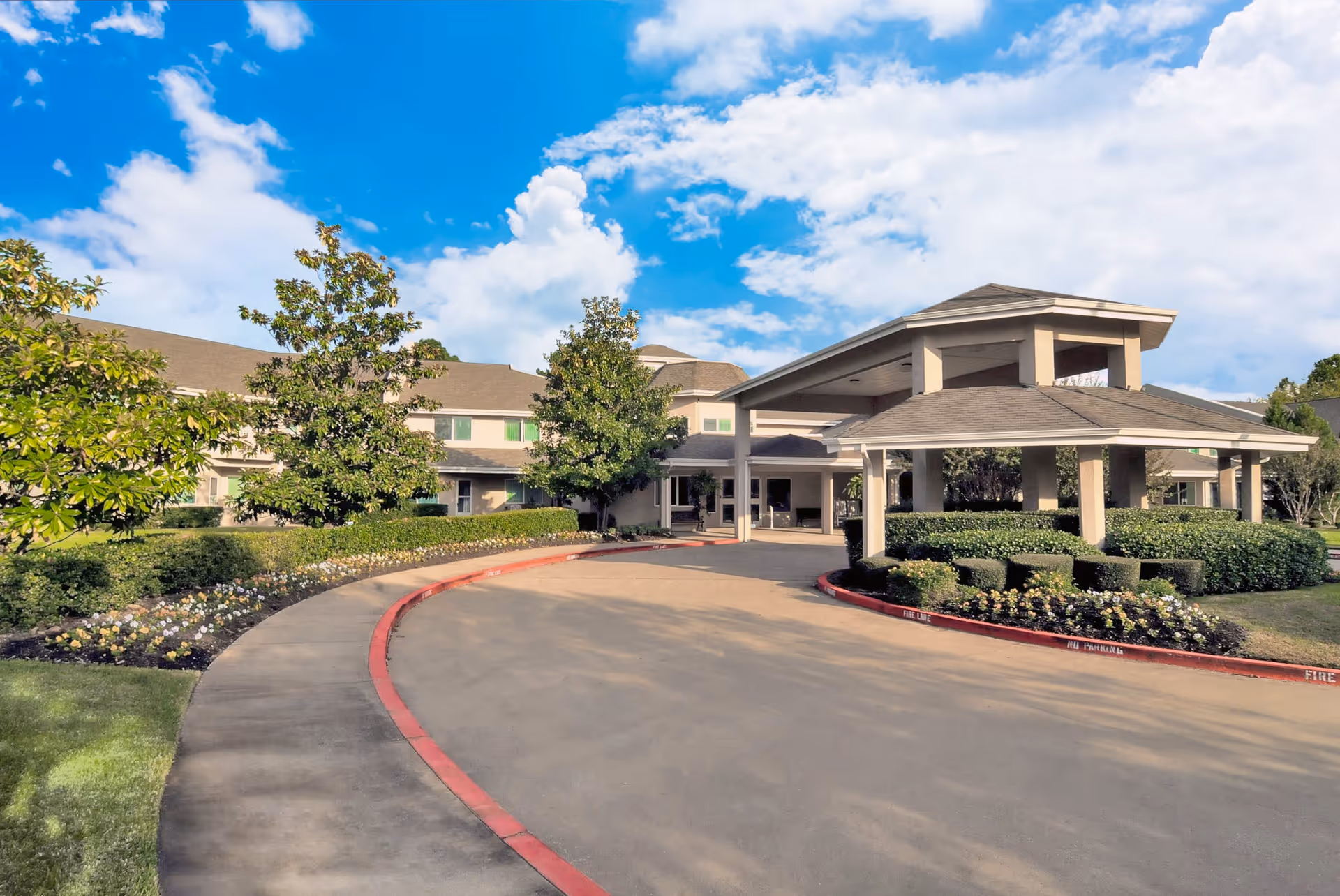 Exterior view of Asher Point Independent Living of Tyler showing a curved driveway leading to a covered entrance with well-maintained landscaping including bushes, flowers, and trees under a partly cloudy blue sky.