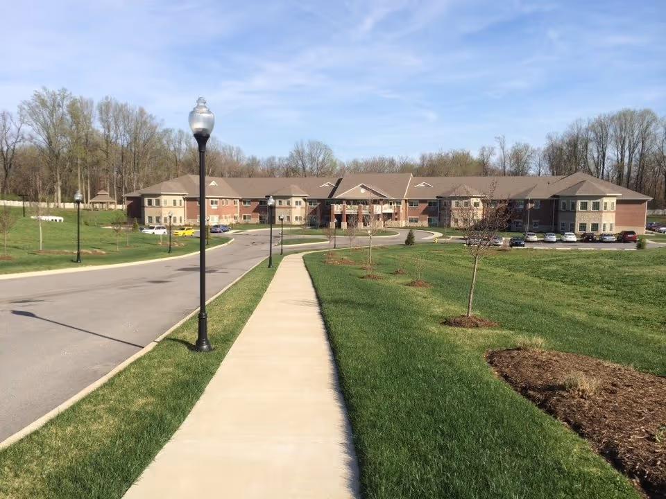 Exterior view of Magnolia Springs East Louisville: a two-story senior living building set behind a sidewalk, lamp posts, driveway, and landscaped lawn.