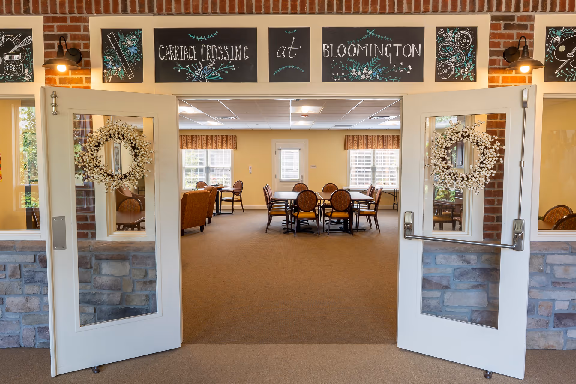Open double doors decorated with white berry wreaths lead into a well-lit common area with tables and chairs. Above the doors are decorative blackboards with floral designs and the text 'Carriage Crossing at Bloomington'. The walls are a warm beige color, and windows with patterned valances allow natural light into the room.