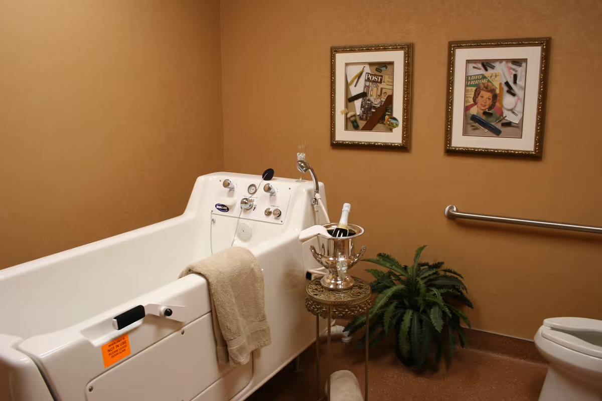 A bathroom with a white walk-in bathtub equipped with various controls and a beige towel hanging on the side. Next to the tub is a small ornate table holding a silver ice bucket with a bottle and a white cloth. There is a green potted plant on the floor beside the table. The walls are painted a warm beige color and decorated with two framed vintage-style pictures. A metal grab bar is mounted on the wall near a white toilet.