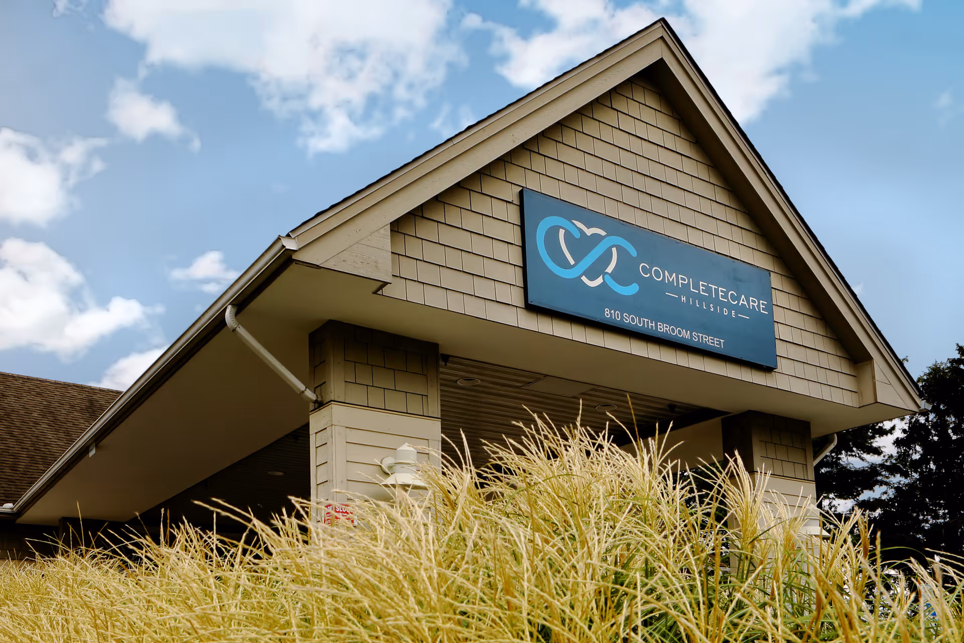 Exterior view of the entrance to Complete Care at Hillside facility with a sign displaying the name and address 810 South Broom Street, under a partly cloudy sky and surrounded by tall ornamental grasses.