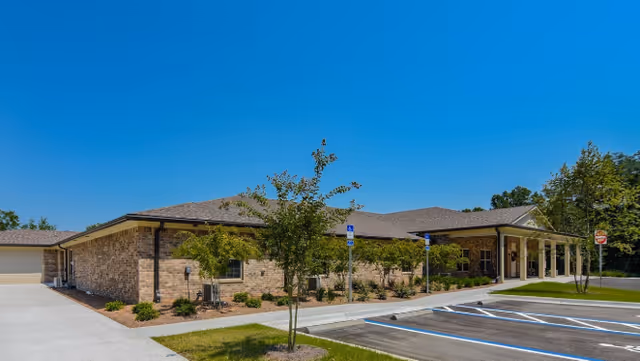 Exterior view of a single-story brick building with a covered entrance, surrounded by small trees and landscaping, under a clear blue sky. There is a parking lot with marked handicap spaces in front of the building.