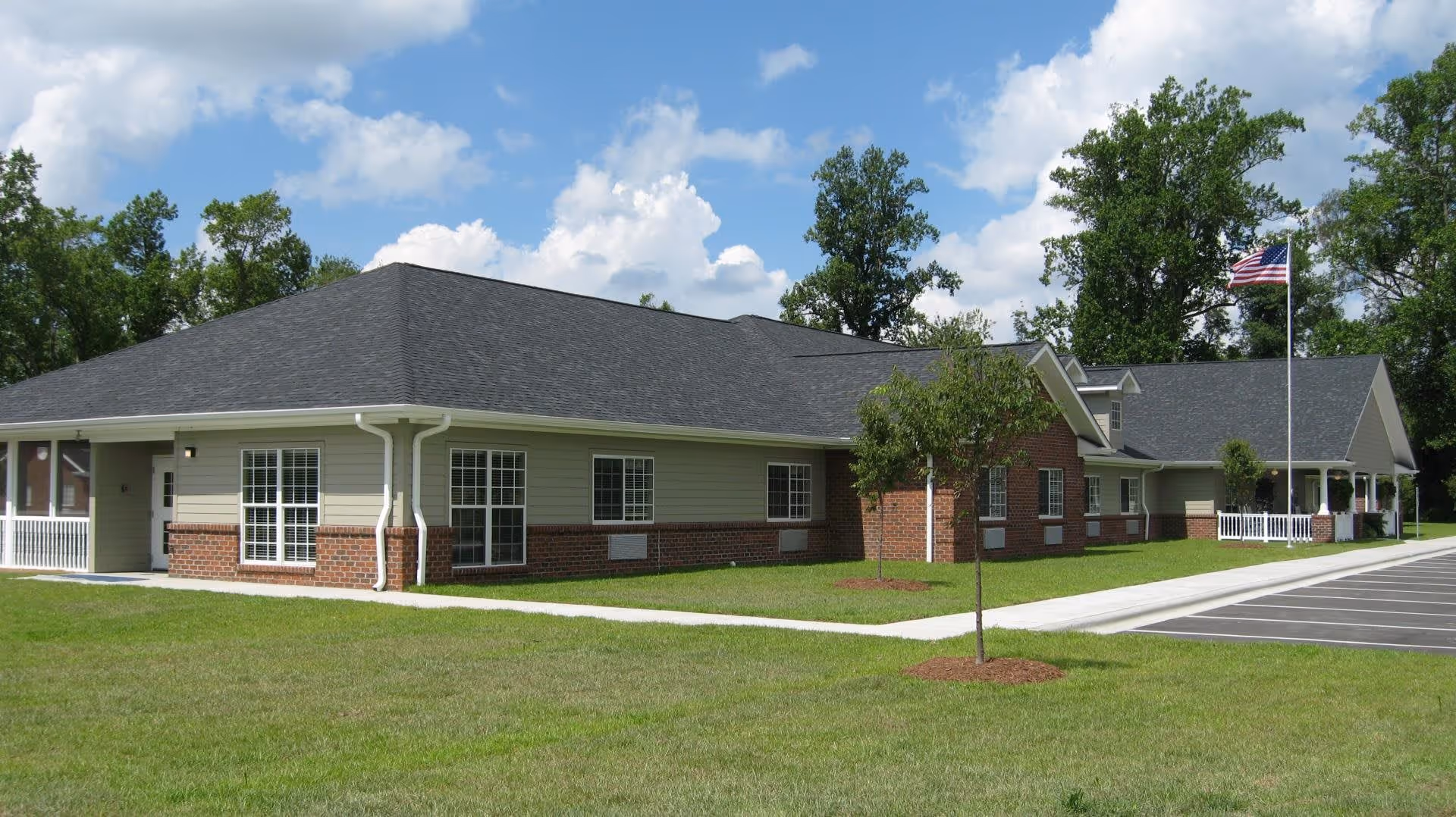 Single-story building with a dark gray roof, beige siding, and red brick accents, surrounded by green grass and trees under a partly cloudy blue sky. There is a paved parking lot to the right and an American flag on a flagpole near the entrance.