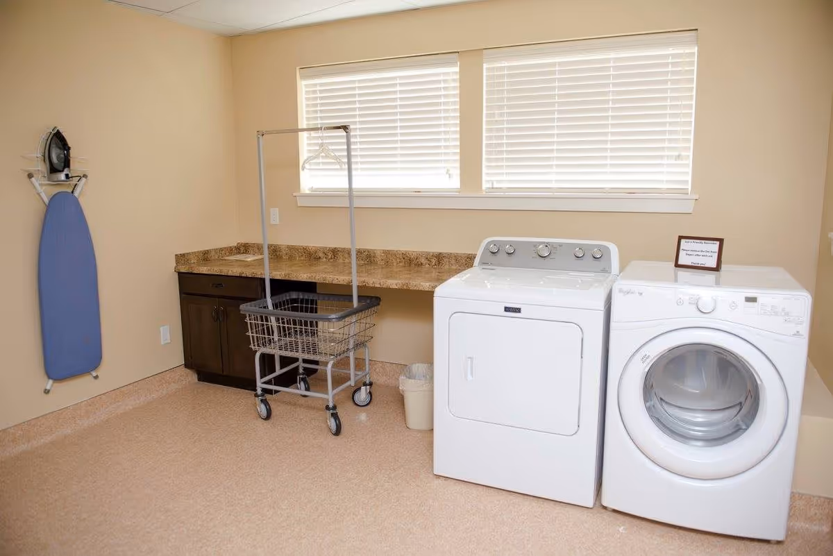 Laundry room with a white washing machine and dryer side by side, a rolling laundry cart with a hanging rod, an ironing board mounted on the wall, and a countertop with cabinets underneath. Two windows with blinds are above the countertop.