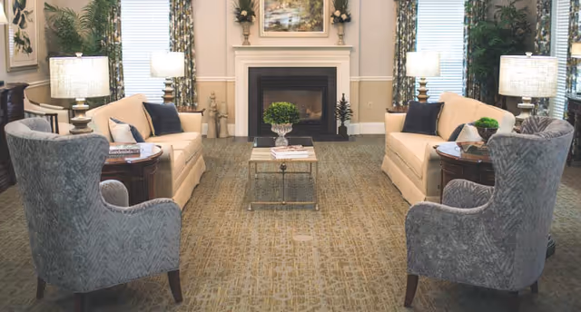 A cozy living room with two beige sofas facing each other, two gray armchairs, a central coffee table with a plant, side tables with lamps, a fireplace with a painting above it, and windows with patterned curtains.