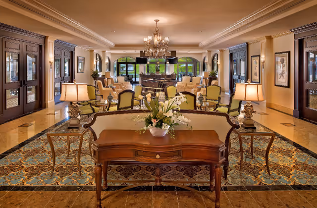Elegant senior living facility common area with classic wooden furniture, floral centerpiece on a wooden table, multiple armchairs arranged around a grand piano, chandeliers hanging from the ceiling, and large windows letting in natural light.