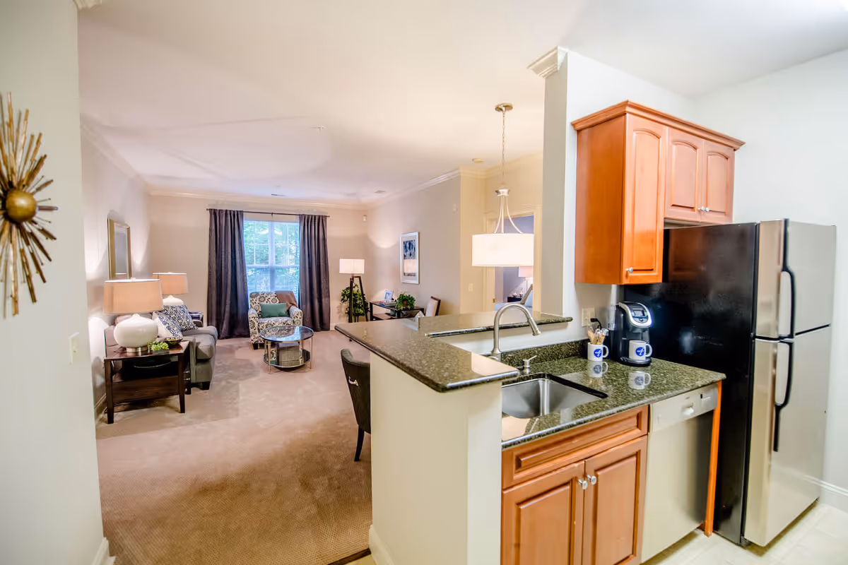 Interior view of a senior living facility apartment showing a kitchen area with wooden cabinets, a black refrigerator, a dishwasher, and a sink with a coffee maker on the counter. The kitchen opens into a living room with a sofa, armchair, coffee table, side tables with lamps, a window with curtains, and a floor lamp near a small desk with a chair and plants.
