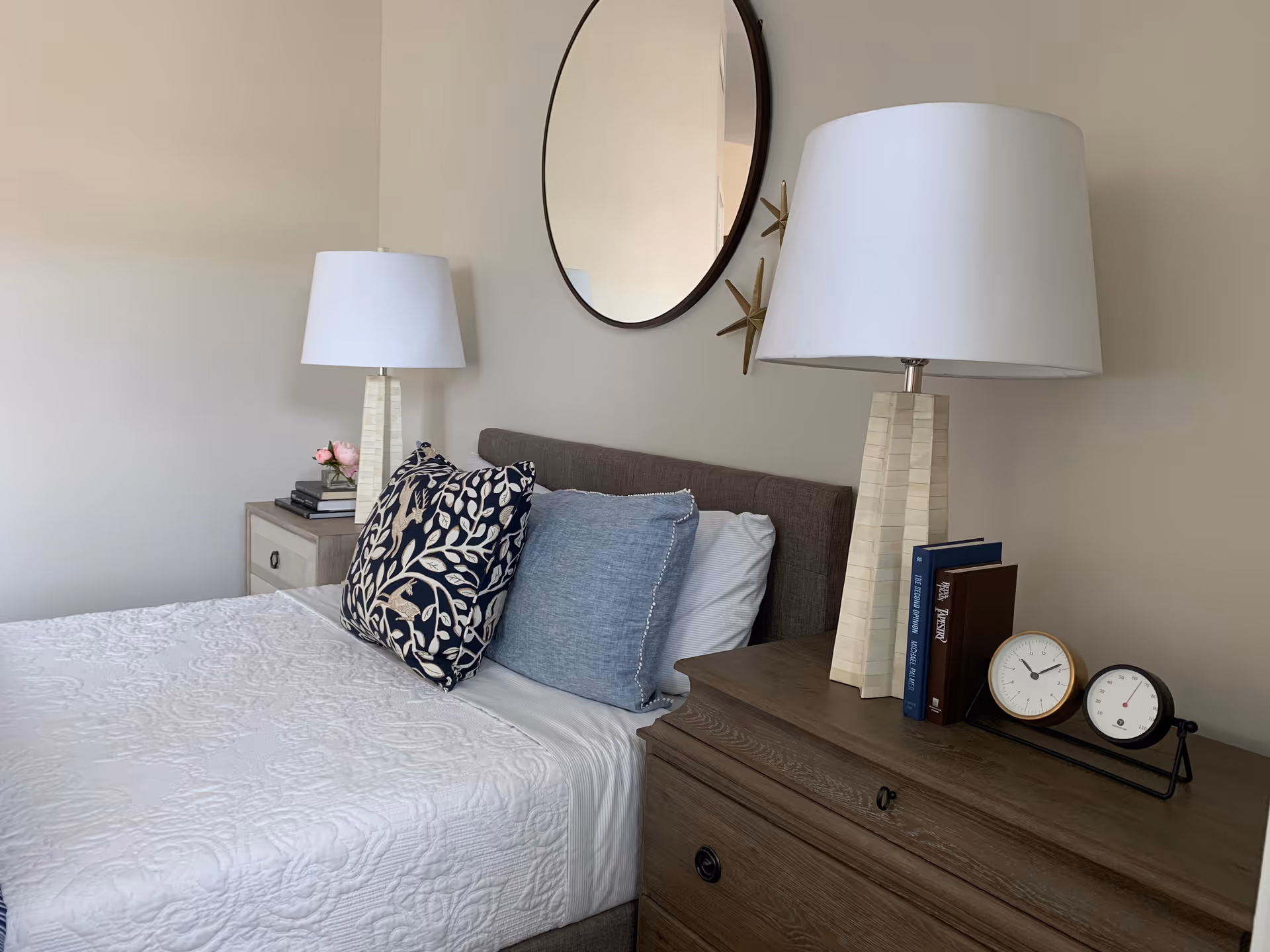 A neatly made bed with a white quilted bedspread and three pillows, two blue and one with a blue and white leaf pattern. On either side of the bed are wooden nightstands with white lamps. One nightstand has a small vase with pink flowers and books, while the other has two clocks and books. A round mirror hangs on the beige wall above the bed.