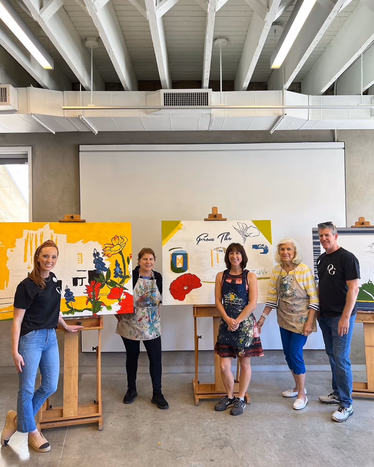 Five adults standing indoors in an art studio with high ceilings and exposed beams. They are posing next to three easels holding colorful paintings in progress. The group includes three women wearing paint-splattered aprons and two people in casual clothing. The atmosphere appears creative and friendly.