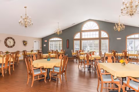 Spacious dining room with multiple round tables covered in yellow tablecloths, wooden chairs, chandeliers, and large arched windows.