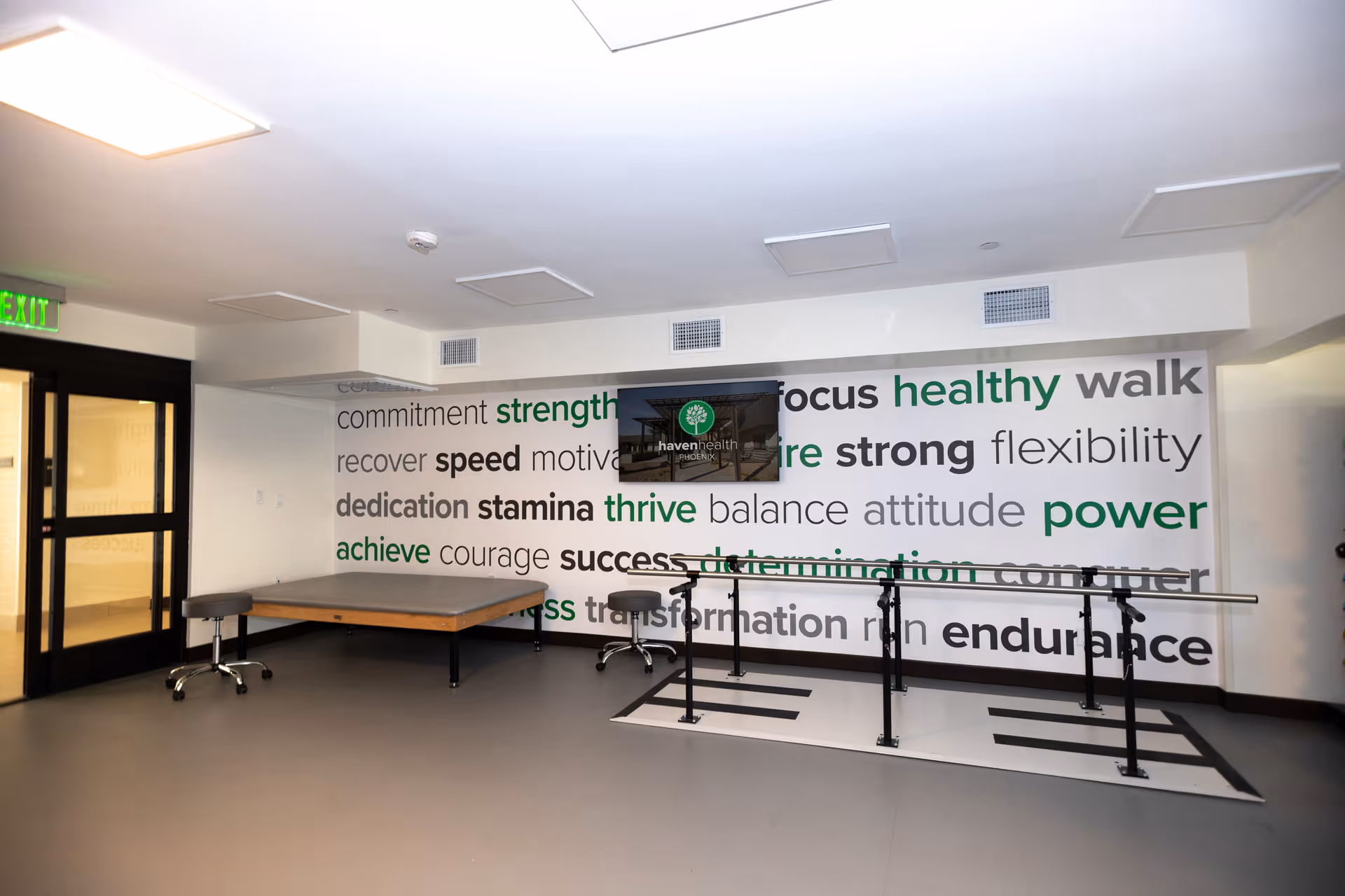 A rehabilitation or physical therapy room with parallel bars for walking exercises, a wooden table, and two stools. The back wall features motivational words such as strength, focus, healthy, power, and endurance in various fonts and colors. A monitor mounted on the wall displays the Haven Health Phoenix logo. There is an exit door on the left side of the room.