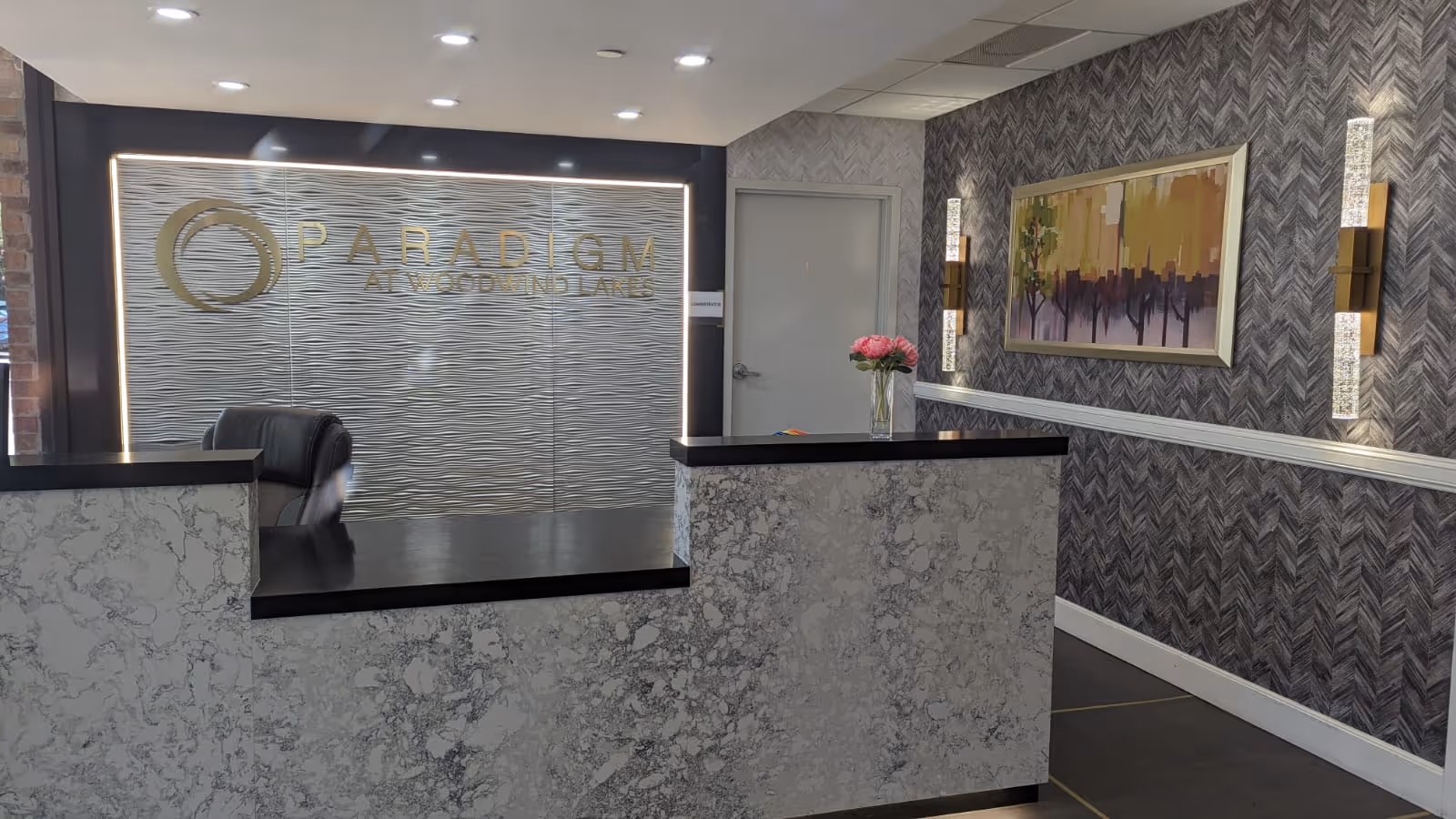 Reception desk and lobby area with a marble-front counter, decorative wallpaper, artwork, and a back wall sign reading "Paradigm at Woodwind Lakes".