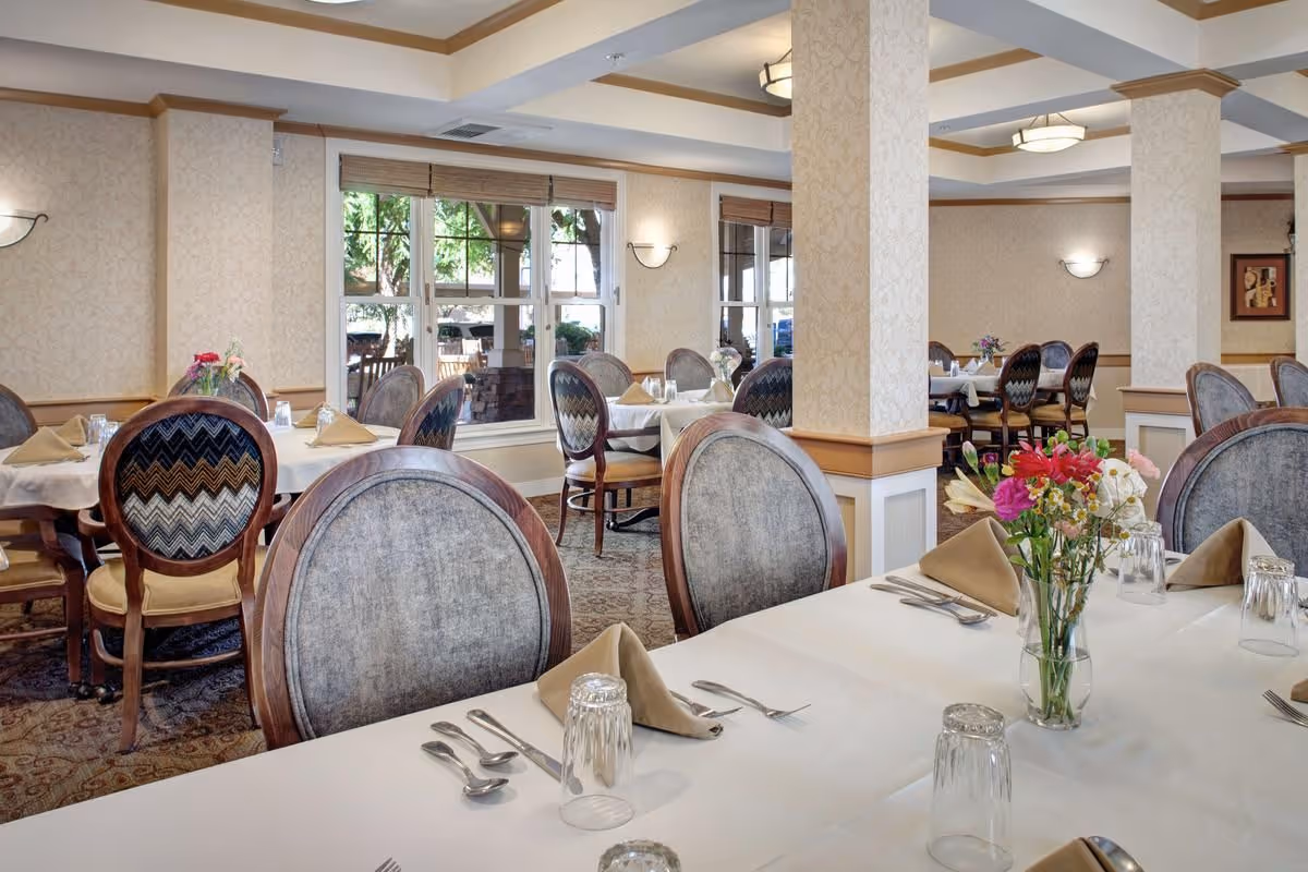 Bright, formal dining room with neatly set tables, upholstered chairs, and floral centerpieces.
