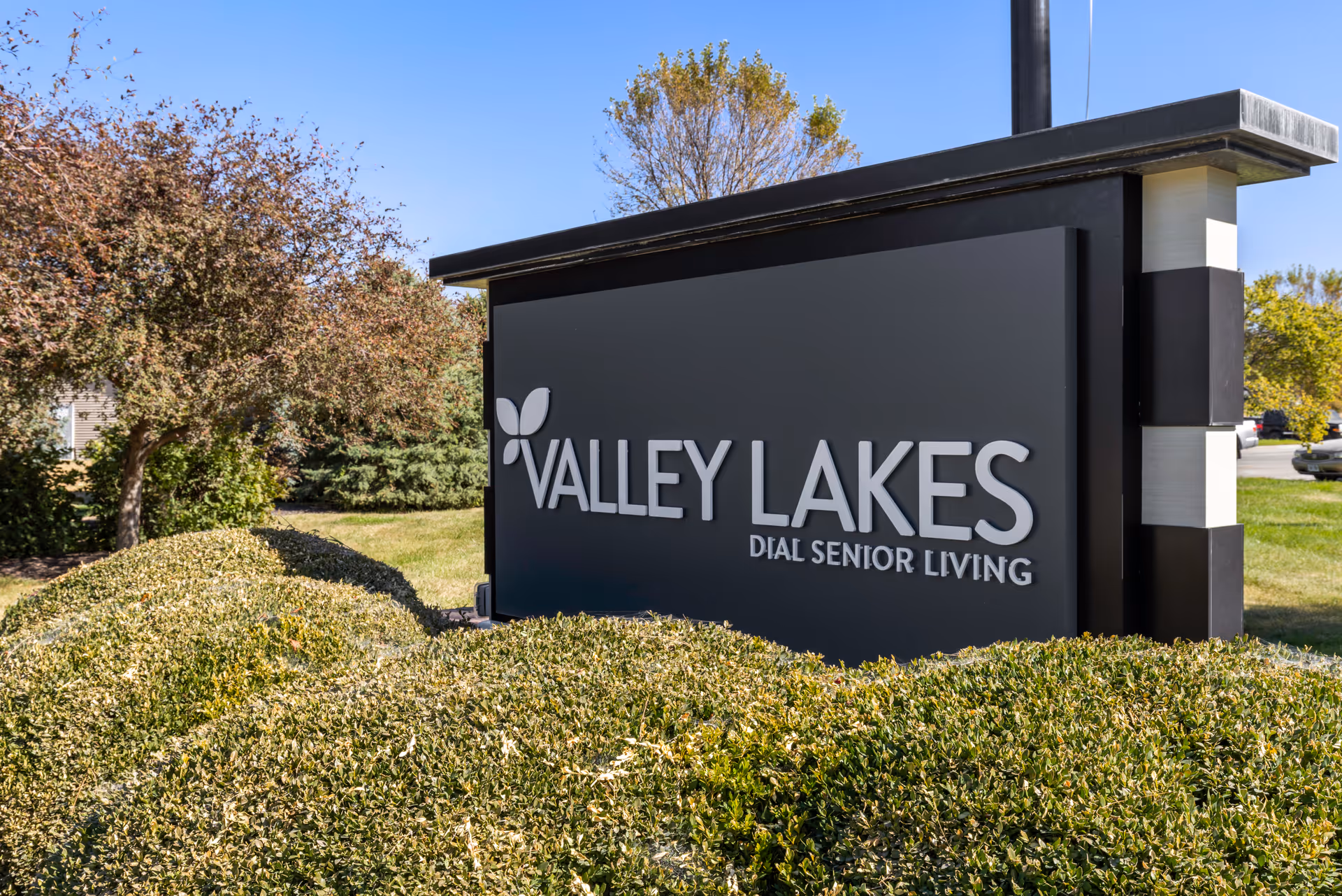 A large outdoor black sign reading "Valley Lakes Dial Senior Living" set among trimmed bushes and trees.