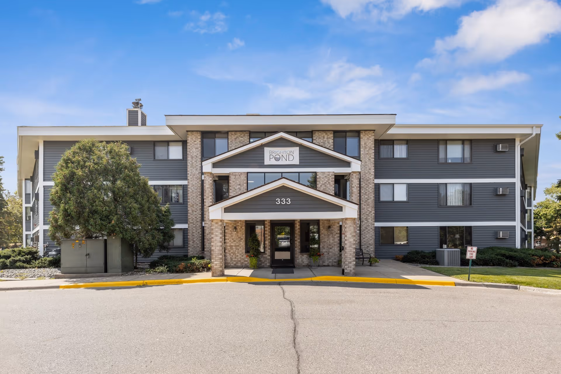 Front exterior view of Brighton Pond Apartments building with a three-story structure, gray siding, brick columns, and a covered entrance with the number 333 above the door. There are trees and shrubs around the building and a clear blue sky in the background.