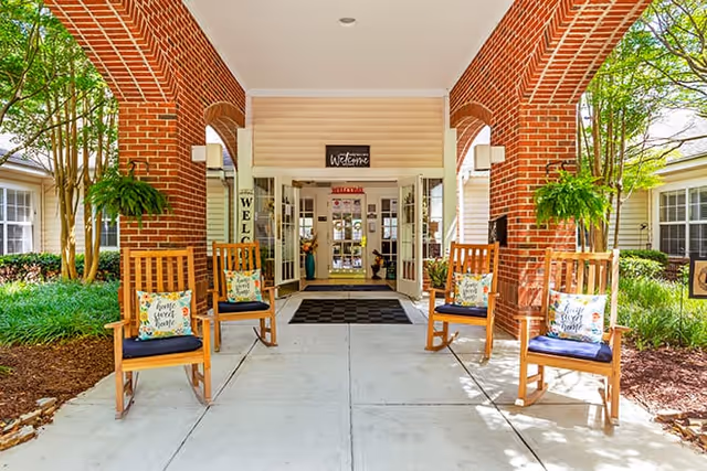 Covered brick-arched entrance with wooden rocking chairs, potted plants, and a welcome sign above double doors.