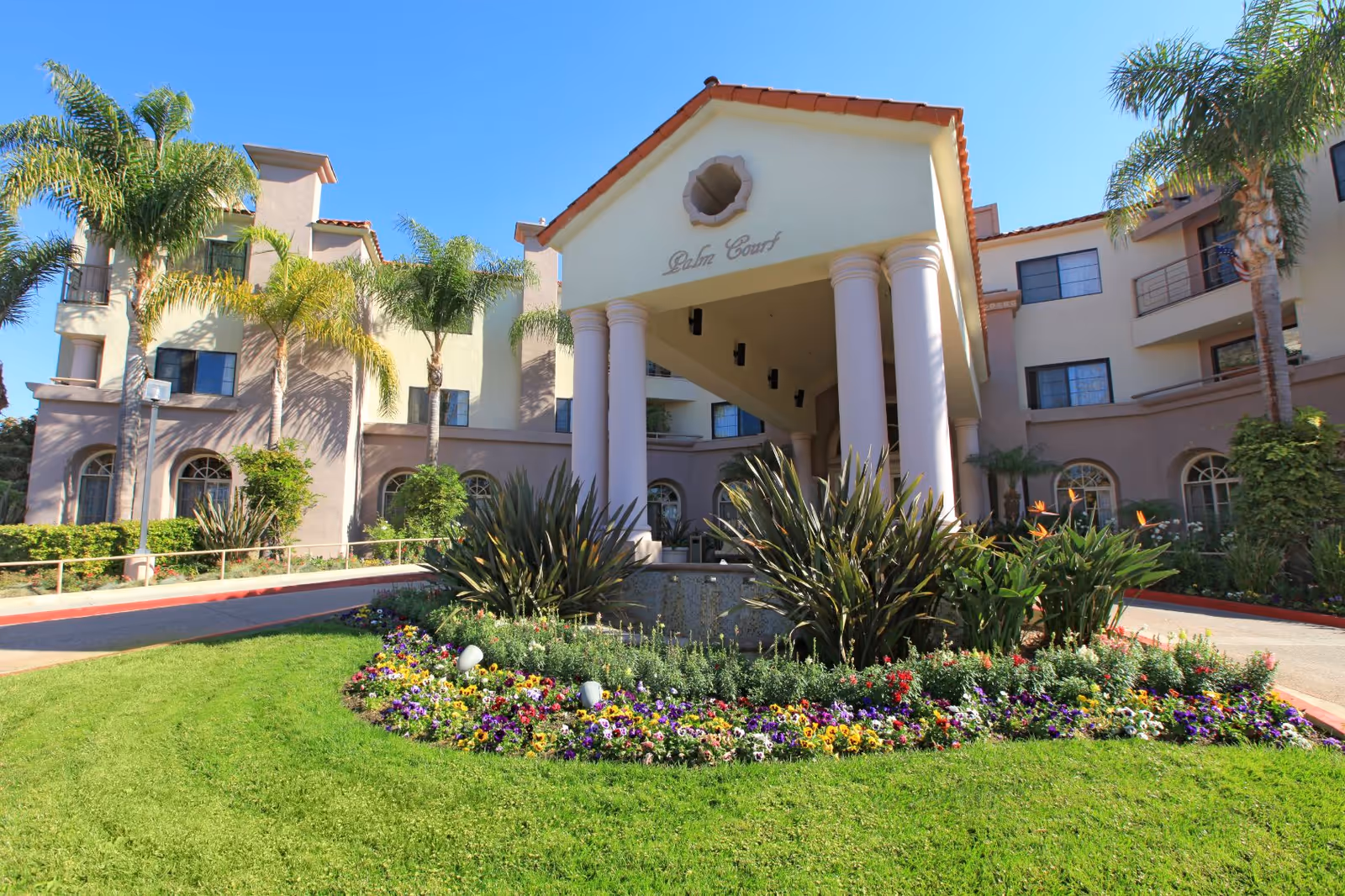 Front exterior view of the Palm Court senior living facility with a covered entrance supported by columns, surrounded by palm trees, green grass, and colorful flower beds under a clear blue sky.