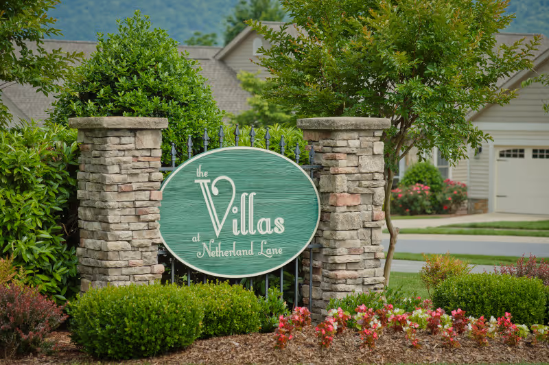 A green oval sign with white text reading 'the Villas at Netherland Lane' mounted on a black iron fence between two stone pillars. The sign is surrounded by neatly trimmed bushes and colorful flowers, with residential houses and trees visible in the background.