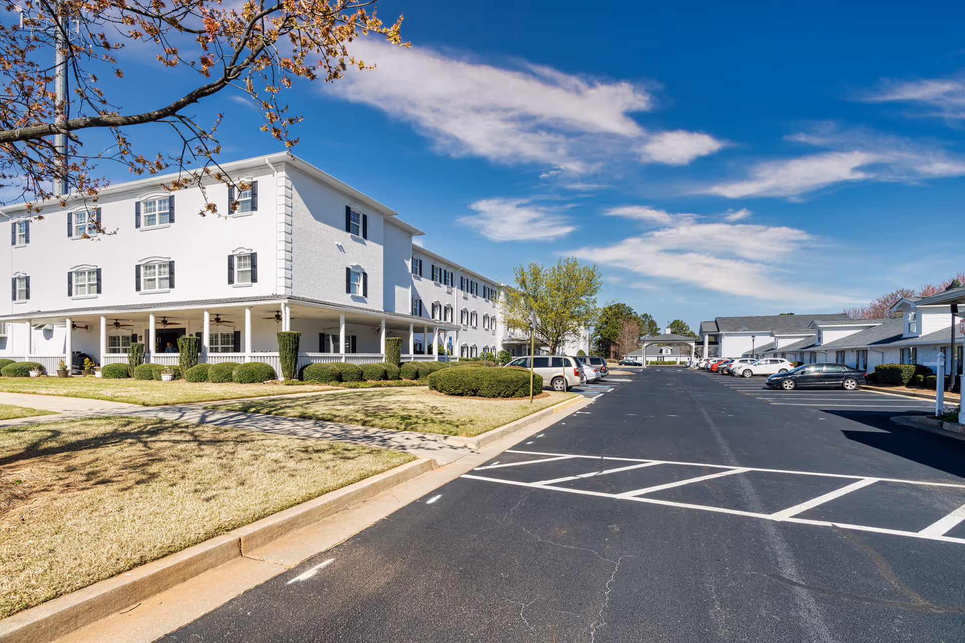 Large white three-story senior living building with a wraparound porch and adjacent parking lot under a blue sky.