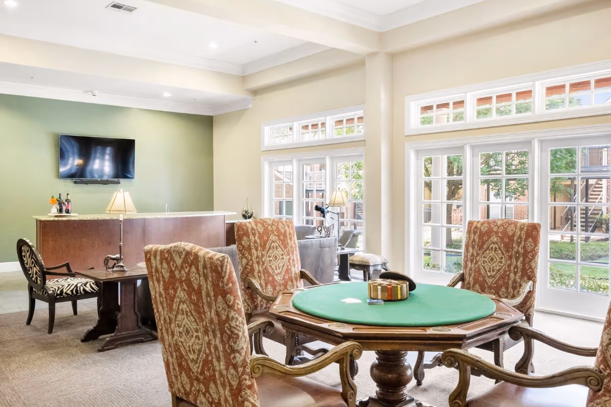 A bright and spacious common area in Preston Place featuring a round poker table with four upholstered chairs, a small bar with a lamp and wine bottles, a wall-mounted TV, and large windows and glass doors letting in natural light and offering a view of the outdoor garden area.