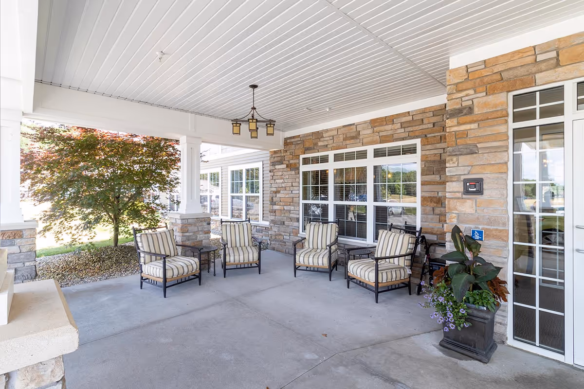 Covered front porch with a stone facade, windows, potted plants, and several cushioned chairs arranged under a hanging light near the entrance.