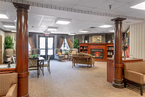 Spacious assisted living common lounge with chairs, tables, decorative columns, a fireplace mantle, and glass entrance doors.