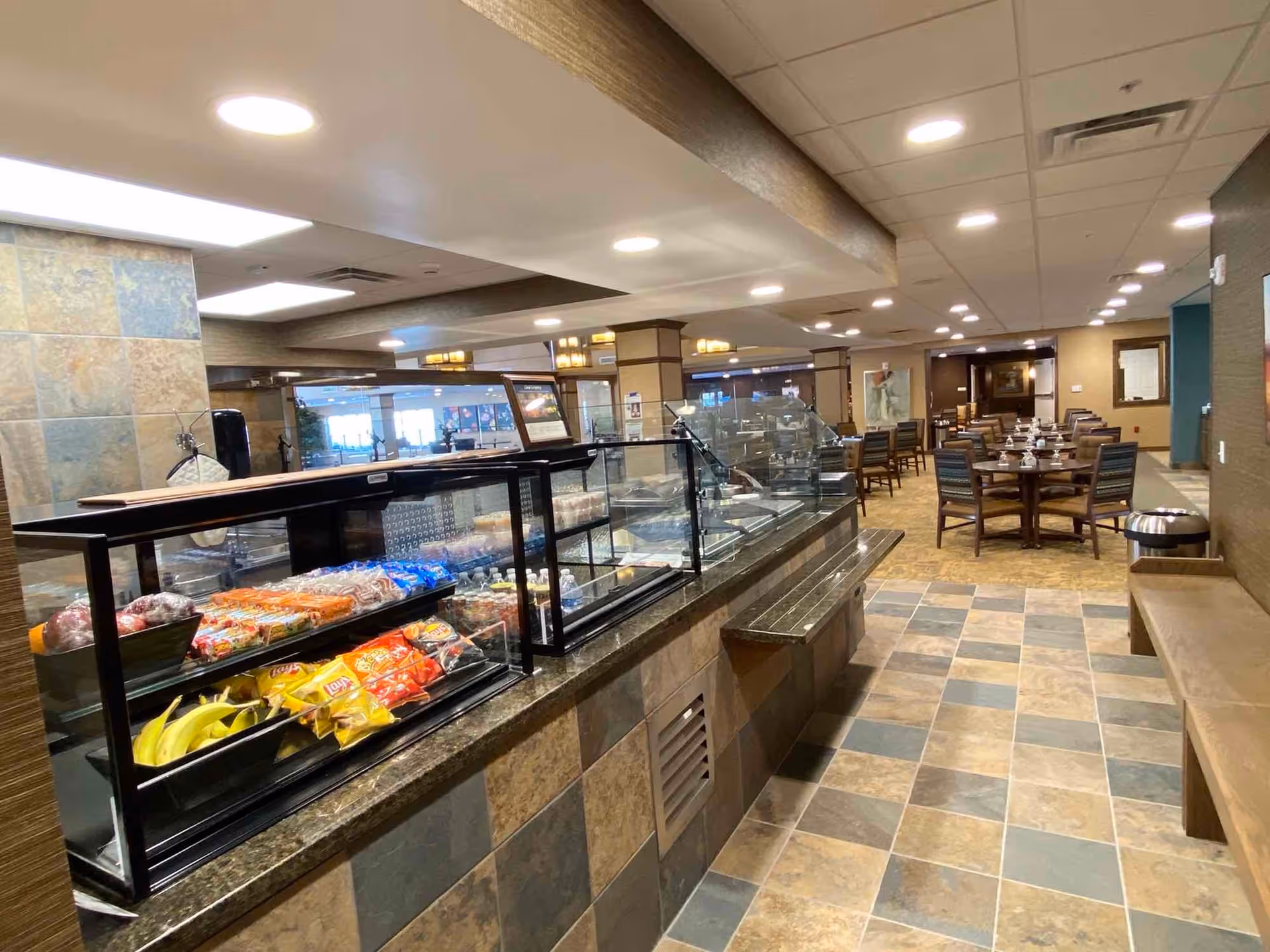 Glass-covered serving counter stocked with snacks and fruit in a dining area with tables and chairs.