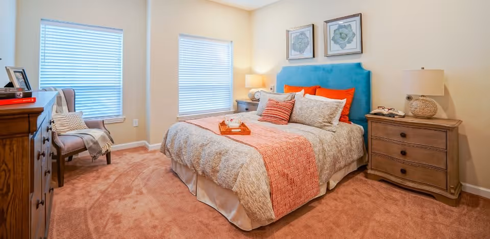A cozy bedroom with a large bed featuring a blue upholstered headboard, beige and orange bedding, and multiple pillows. There are two framed botanical prints on the wall above the bed. On either side of the bed are wooden nightstands with lamps. A wooden dresser with books and a framed photo is on the left side, and a cushioned chair with a throw blanket is positioned near two windows with blinds. The room has beige walls and carpeted flooring.
