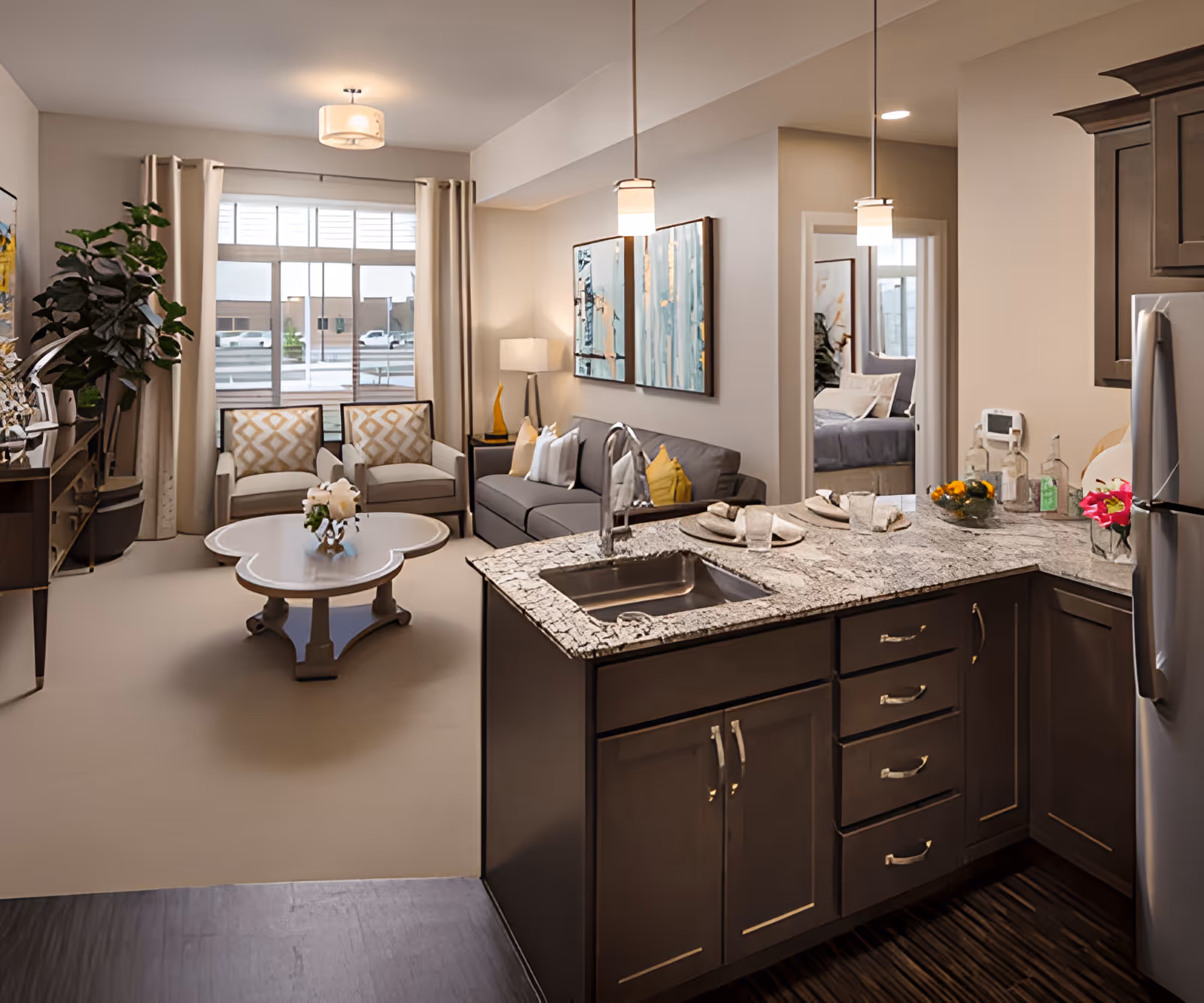 A bright and modern assisted living apartment featuring an open kitchen with dark wood cabinets, a granite countertop with a sink, and stainless steel refrigerator. The kitchen opens to a cozy living area with a gray sofa, two patterned armchairs, a white coffee table with flowers, and large windows with beige curtains. A bedroom is visible through an open doorway, and the space is decorated with contemporary artwork and pendant lighting.