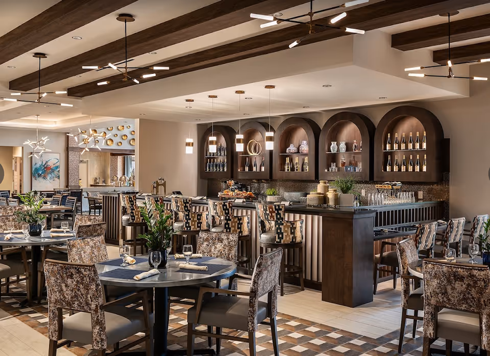 A modern dining area with multiple round tables and chairs, a bar with high stools, decorative shelves with bottles and ornaments, and stylish ceiling lights with wooden beams.