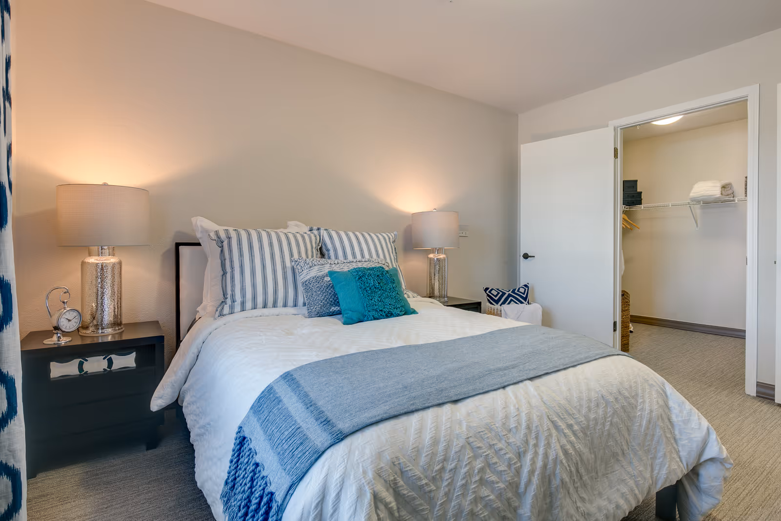A neatly made bed with white and blue bedding and multiple pillows in a bedroom. There are two bedside tables with lamps on each side of the bed. An open door reveals a walk-in closet with shelves and hanging space. The room has light-colored walls and carpeted flooring.