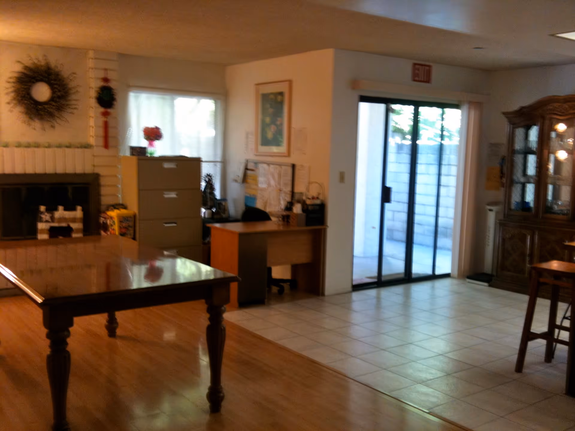 Interior view of a senior living facility room with a wooden table in the foreground, a fireplace with decorative items on the left wall, a filing cabinet and desk with office supplies near a window, and a glass sliding door leading outside. The floor transitions from wood to tile, and there is a wooden cabinet with glass doors on the right side.