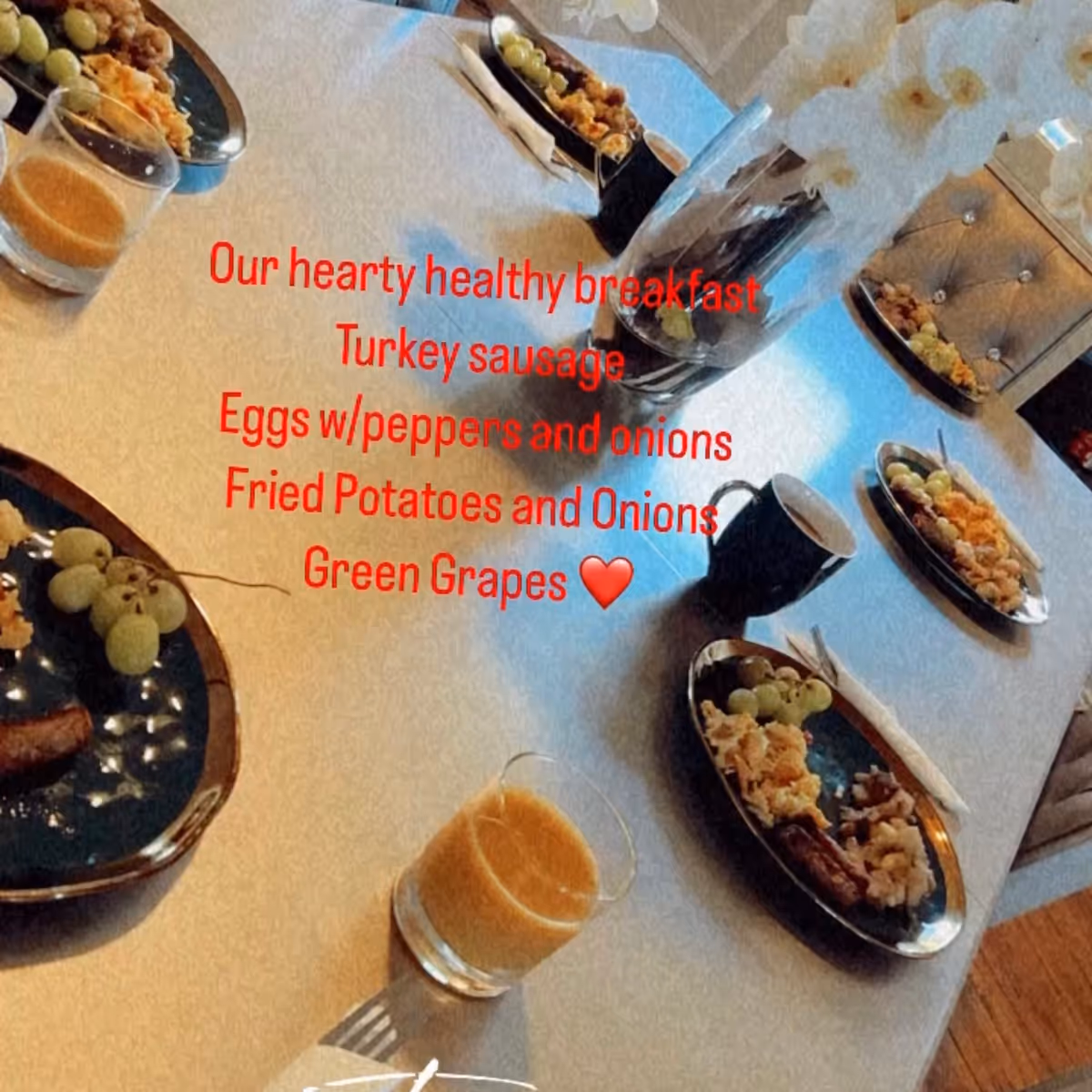 A dining table set with plates of breakfast food including turkey sausage, scrambled eggs with peppers and onions, fried potatoes with onions, and green grapes. There are also glasses of orange juice and coffee cups on the table. A vase with white flowers is placed in the center of the table. Red text overlay describes the meal.