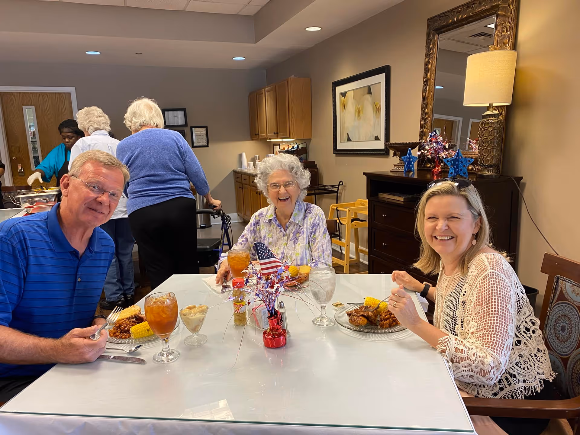 Three people sitting at a dining table in a senior living facility, smiling and enjoying a meal with iced tea and desserts. The table is decorated with a small patriotic centerpiece featuring an American flag. In the background, other people are serving food from a buffet in a room with beige walls, wooden cabinets, and framed artwork.