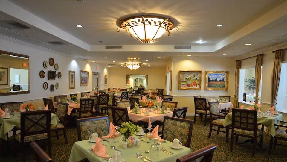 A well-lit dining room with multiple tables covered in pastel green and pink tablecloths, each set with cups, glasses, silverware, and folded napkins. The chairs have floral upholstery, and the walls are decorated with framed paintings and a mirror. Large windows with curtains allow natural light into the room.