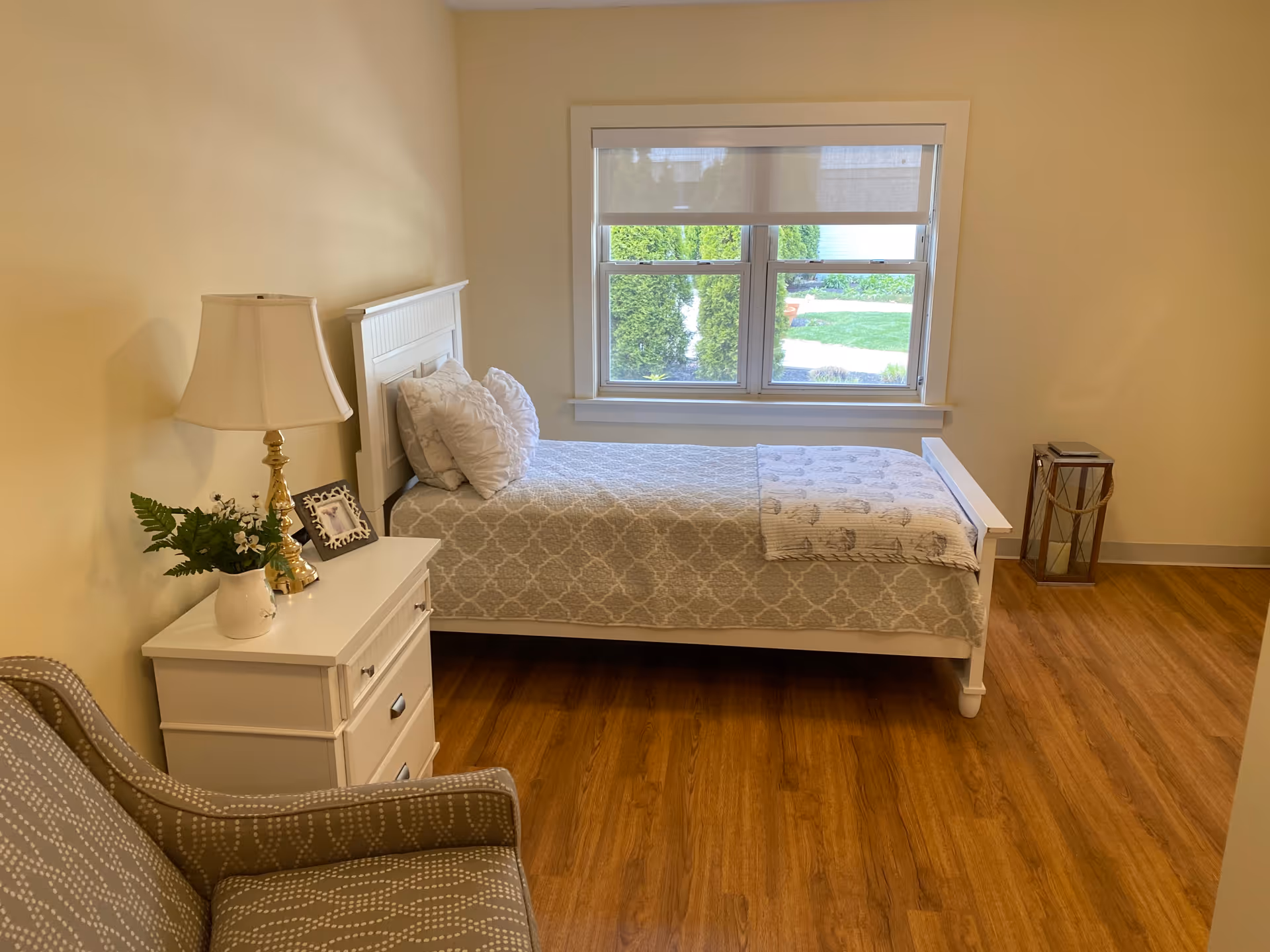 Bright, simply furnished bedroom with a single bed, nightstand with lamp and a chair beside a window.