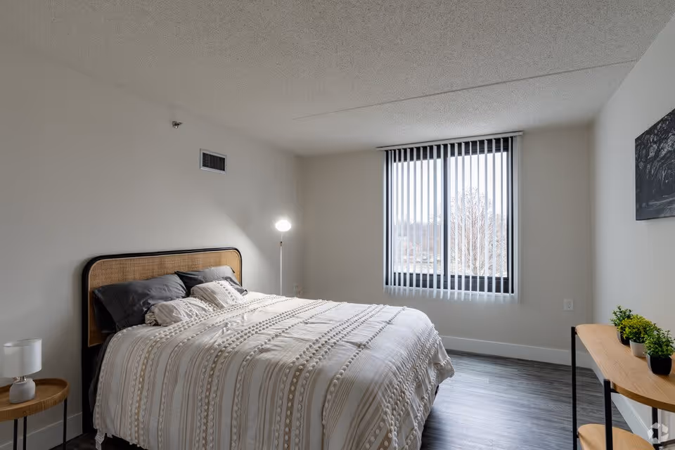 A bright bedroom with a large bed covered in a white and beige patterned comforter and multiple pillows. The room has light-colored walls, a window with vertical blinds letting in natural light, a small round wooden side table with a white lamp, a standing floor lamp, and a wooden console table with small potted plants. The floor is a dark wood laminate.