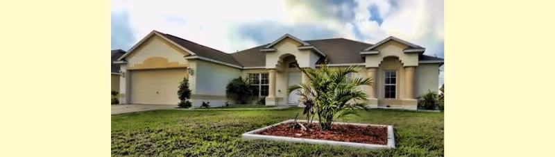 Single-story residential building with a beige exterior, a two-car garage, and a small landscaped area with a palm plant in front under a partly cloudy sky.