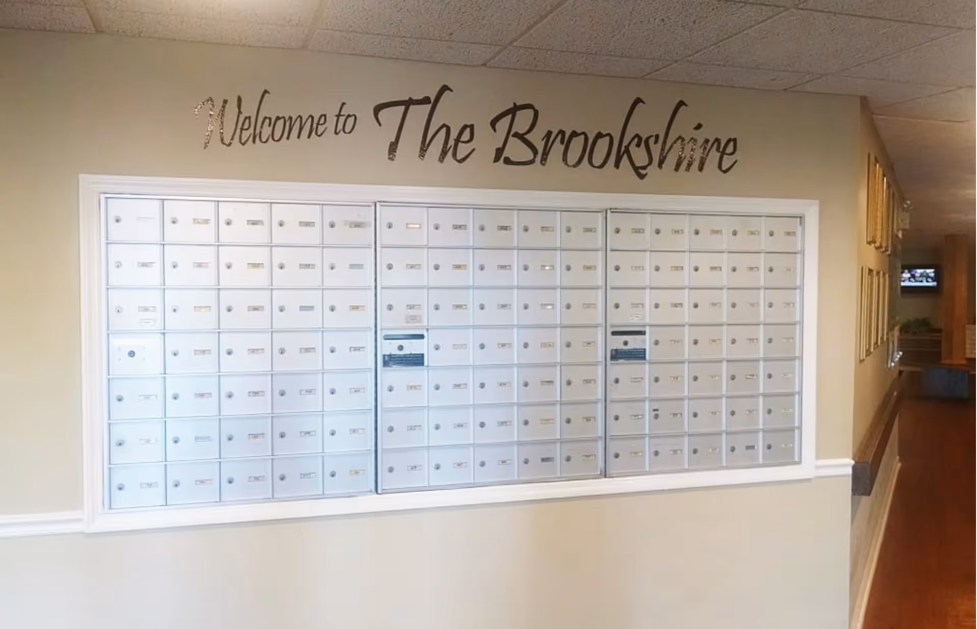 A wall with multiple small mailboxes arranged in a grid, each with a lock and label. Above the mailboxes, the wall has a decorative text that reads 'Welcome to The Brookshire'. The hallway adjacent to the mailboxes has wooden flooring and framed pictures on the wall.