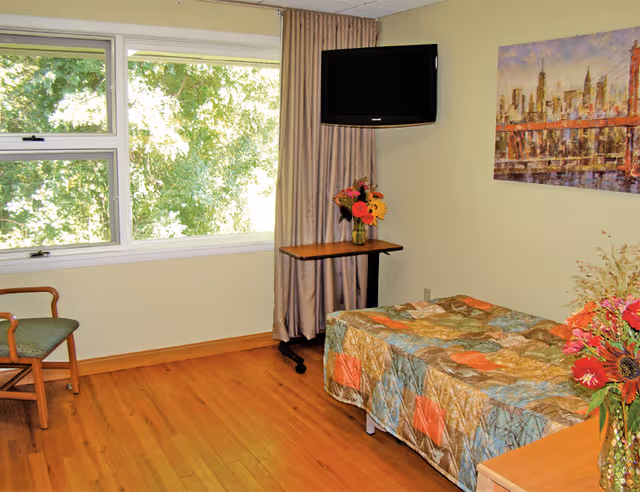 A small bedroom with a single bed covered in a colorful patchwork quilt. There is a wooden chair with a green cushion near a large window showing green trees outside. A small table with a vase of flowers is placed under a wall-mounted flat screen TV. A painting of a cityscape hangs on the wall above the bed. The room has wooden flooring and light yellow walls.