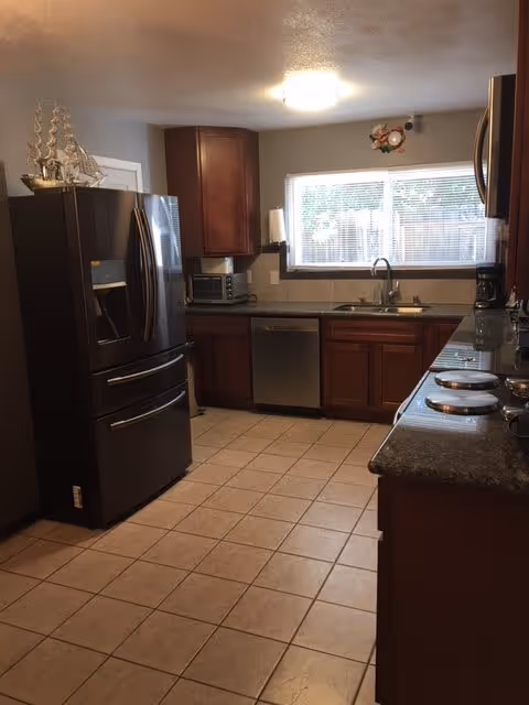 A kitchen with tiled floor, dark wooden cabinets, and granite countertops. There is a stainless steel refrigerator on the left, a dishwasher under the counter, a microwave on the counter, a coffee maker near the window, and a stove with four burners on the right. A window above the sink lets in natural light.