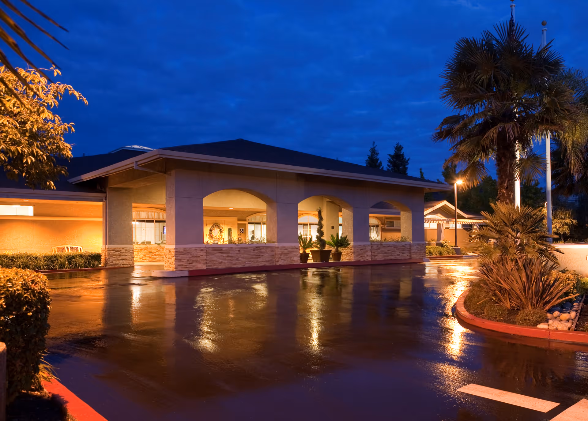 Exterior view of a senior living facility building at dusk with wet pavement reflecting lights, palm trees, and landscaped plants near the entrance.