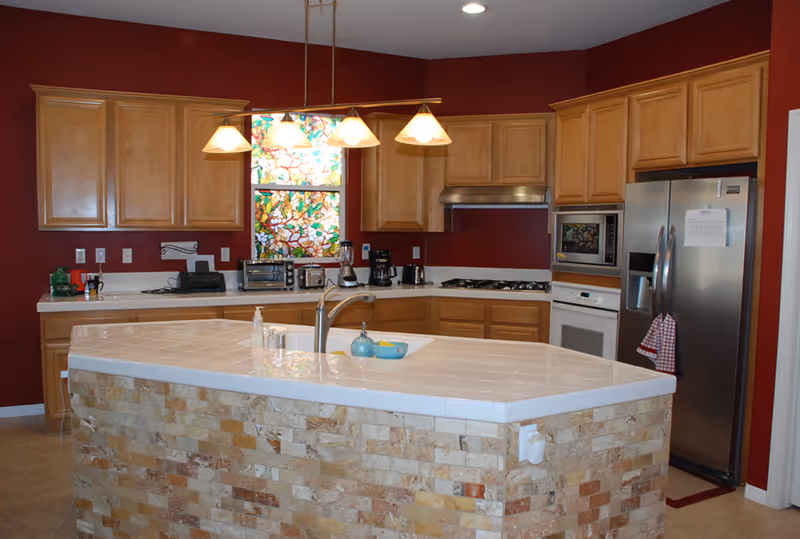 A modern kitchen with wooden cabinets, a stainless steel refrigerator, microwave, oven, and a gas stove. The kitchen island has a tiled countertop with a sink and soap dispenser. There is a stained glass window above the counter and a hanging light fixture with four lamps.