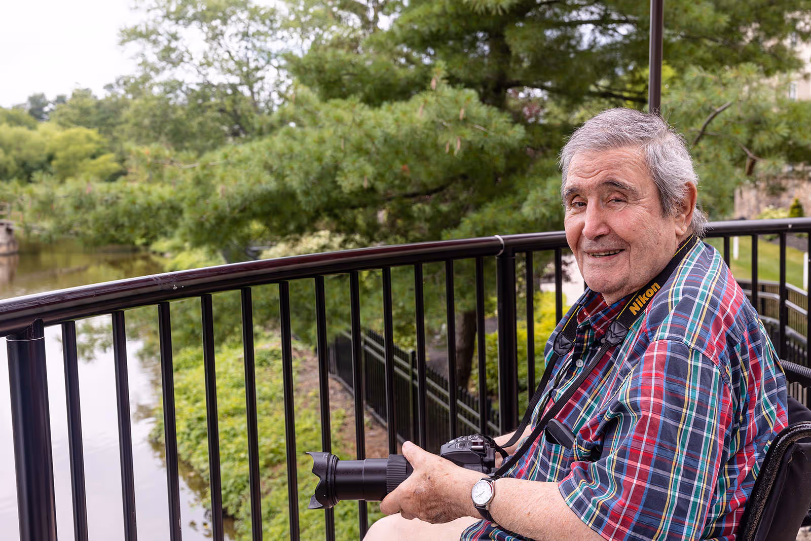 An elderly man sitting in a wheelchair on a balcony or outdoor patio area, holding a Nikon camera with a long lens. He is wearing a colorful plaid shirt and smiling, with green trees and a body of water visible in the background.